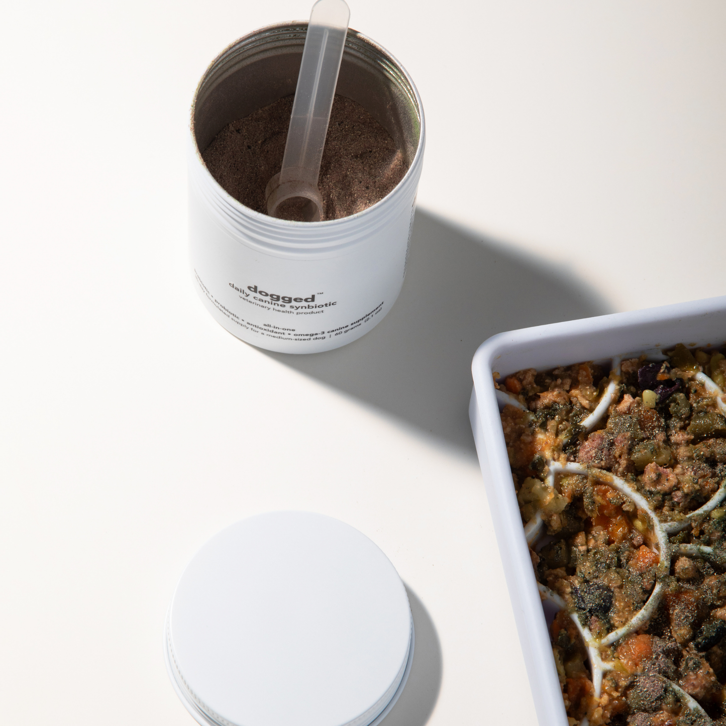 Container of dog food with a scoop next to a dish of cooked food on a white surface